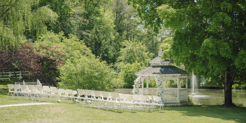 Elegant outdoor wedding ceremony setup with white chairs, gazebo, and lush green trees by a serene pond.