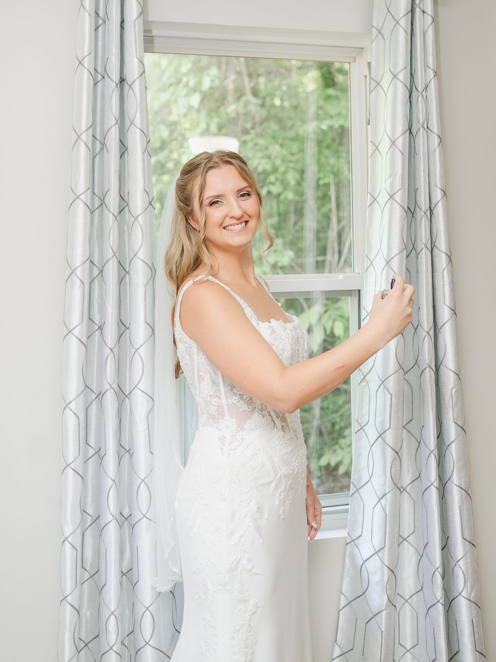 Smiling bride in white lace wedding gown standing by a window with elegant curtains.