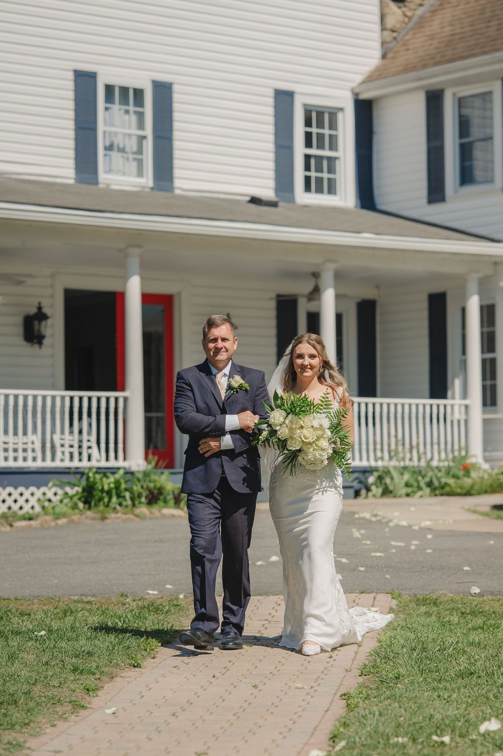 Bride in white gown holding bouquet walks with suited man outside white colonial house with blue shutters.