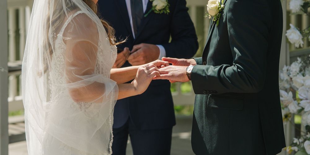 Bride and groom exchanging rings during outdoor wedding ceremony with officiant in background.