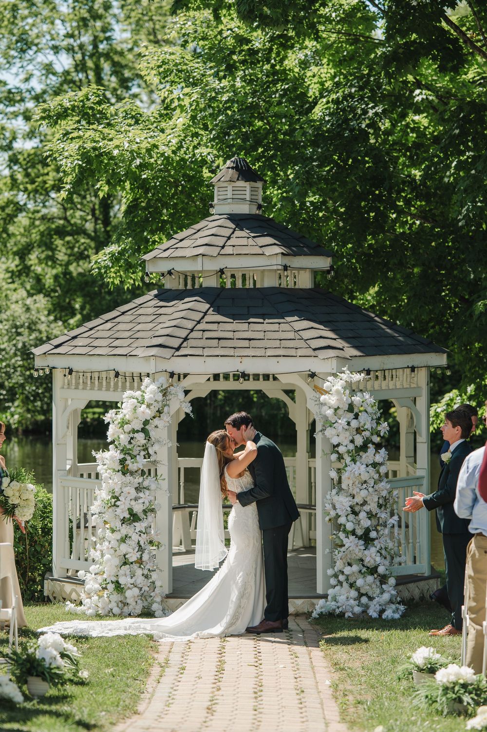 Bride and groom share first kiss at white gazebo altar adorned with lush white floral arrangements during outdoor wedding ceremony.