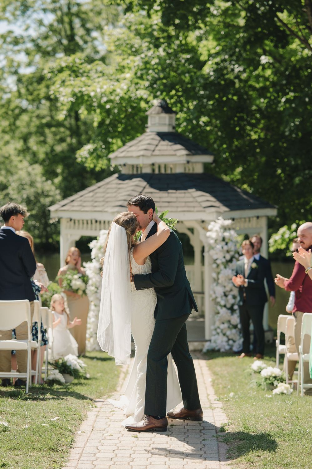 Bride and groom share first kiss as newlyweds on outdoor wedding aisle with white gazebo and floral arch backdrop.