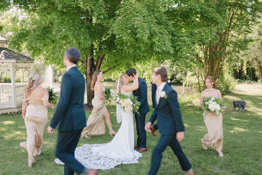Wedding party walks outdoors as bride and groom share a kiss on green lawn near white gazebo.