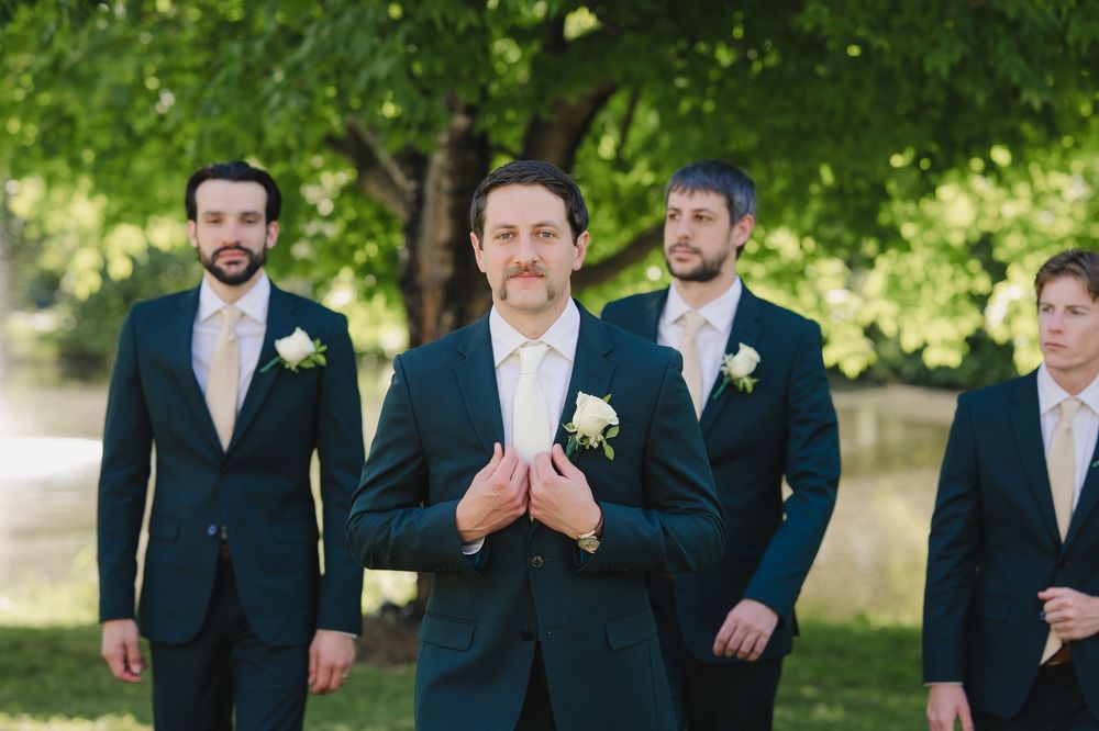 Groom in navy suit clasps hands outdoors with groomsmen wearing matching suits and white boutonnieres at wedding.