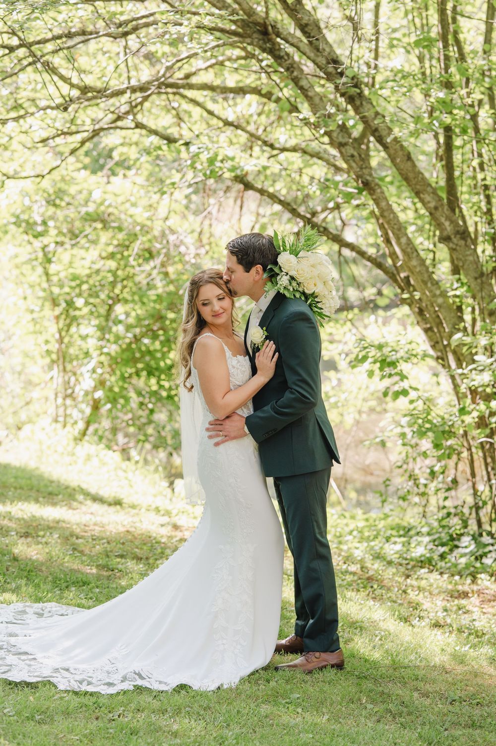 Bride and groom embrace outdoors under lush green trees, bride in lace gown with veil, groom in dark green suit.