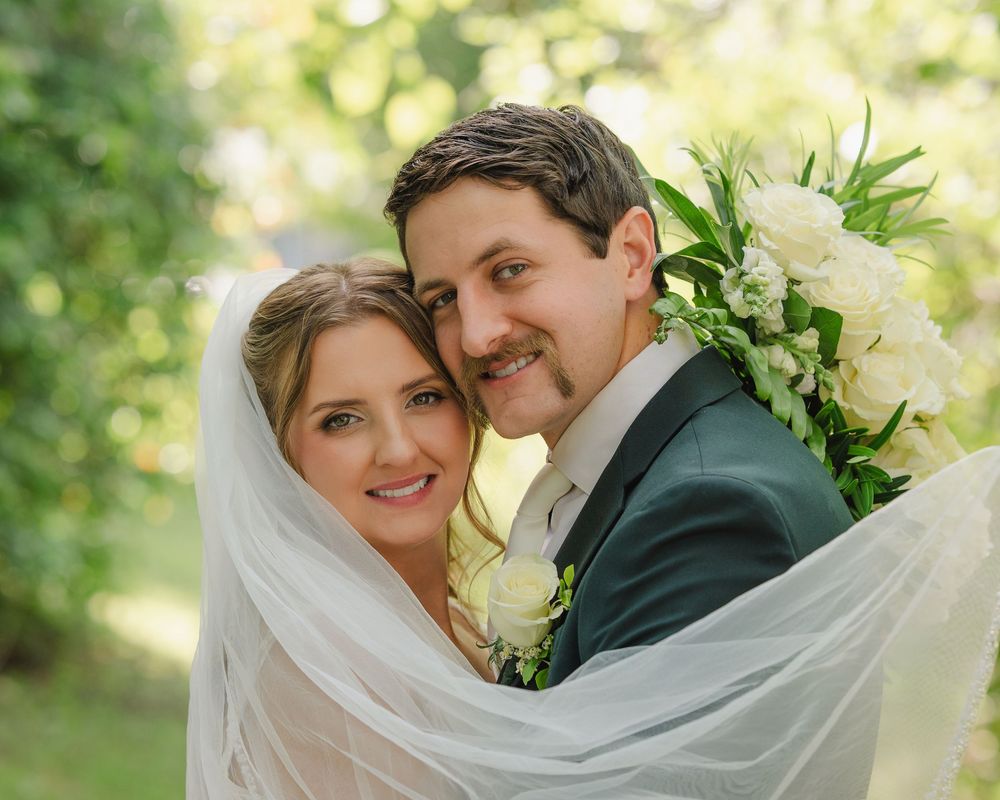 Smiling bride and groom wrapped in veil holding white bouquet in sunlit garden setting.