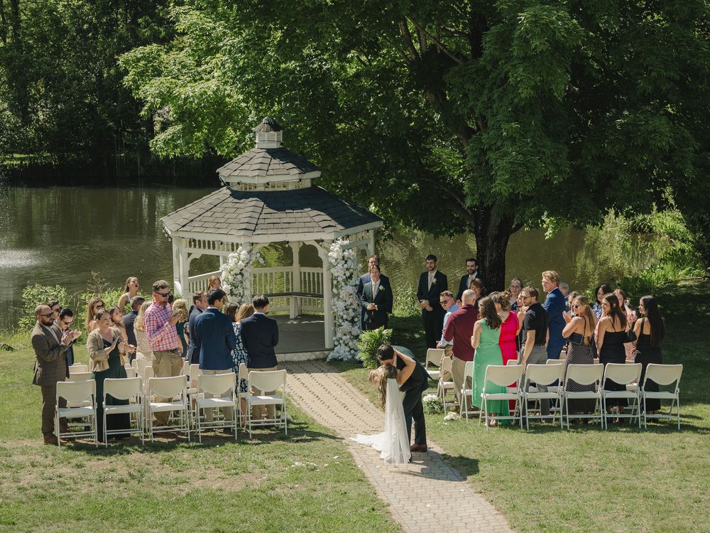 Outdoor wedding ceremony by a gazebo near a pond, with guests seated on white chairs surrounded by lush green trees.