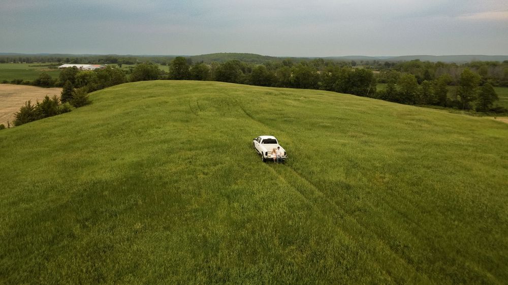 Aerial view of a white vintage car parked alone in a vast green rolling meadow under a cloudy sky.