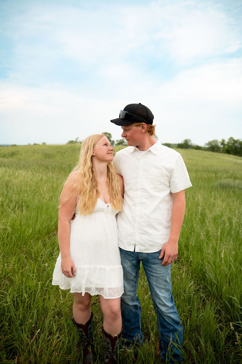 A young couple stands in a lush green field, woman in white dress and boots, man in white shirt and black cap.