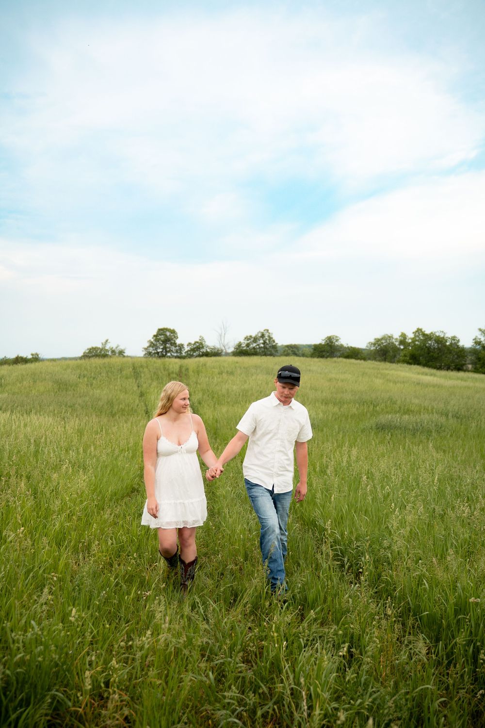 Couple holding hands walking through a lush green field, woman in white dress and man in jeans and cap.