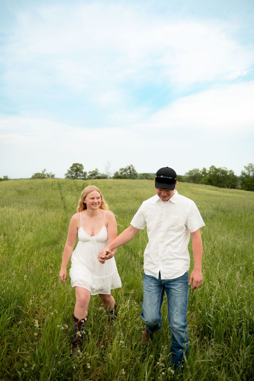 Couple holding hands walking through a lush green field, woman in white dress and cowboy boots, man in white shirt and cap.