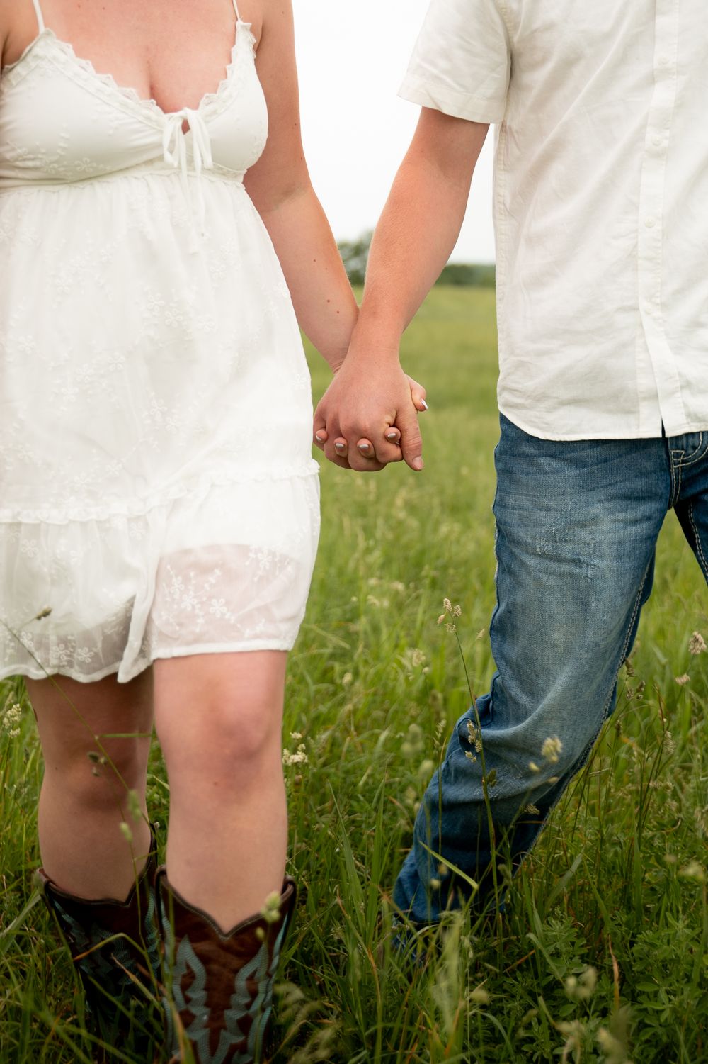 Couple holding hands in a green field, woman wearing white dress with cowboy boots and man in jeans.
