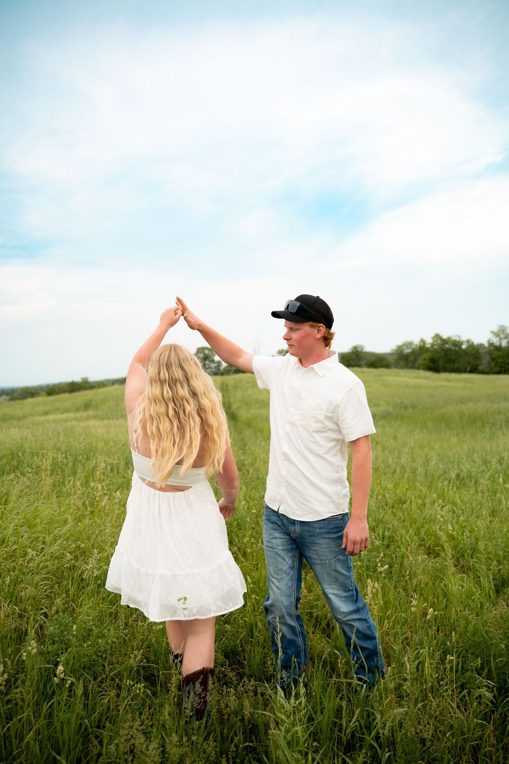 Man in black cap spinning woman in white dress and cowboy boots in a green meadow under a cloudy sky.
