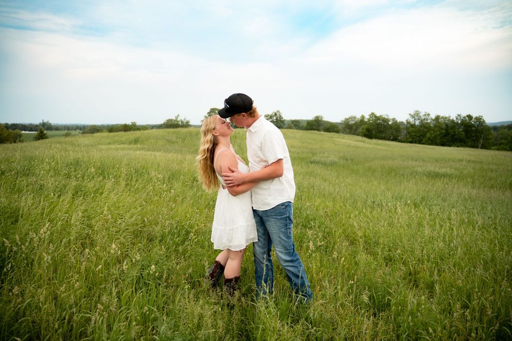 Couple sharing a romantic kiss in a lush green field, woman in white dress and man in jeans and black cap.