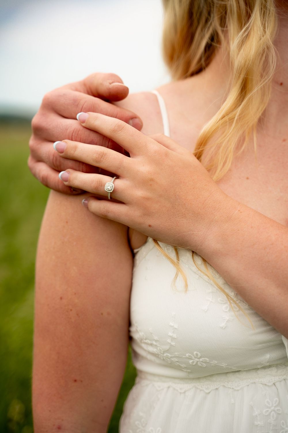 Close-up of an engagement ring on a woman's hand resting on her shoulder in a white dress outdoors.