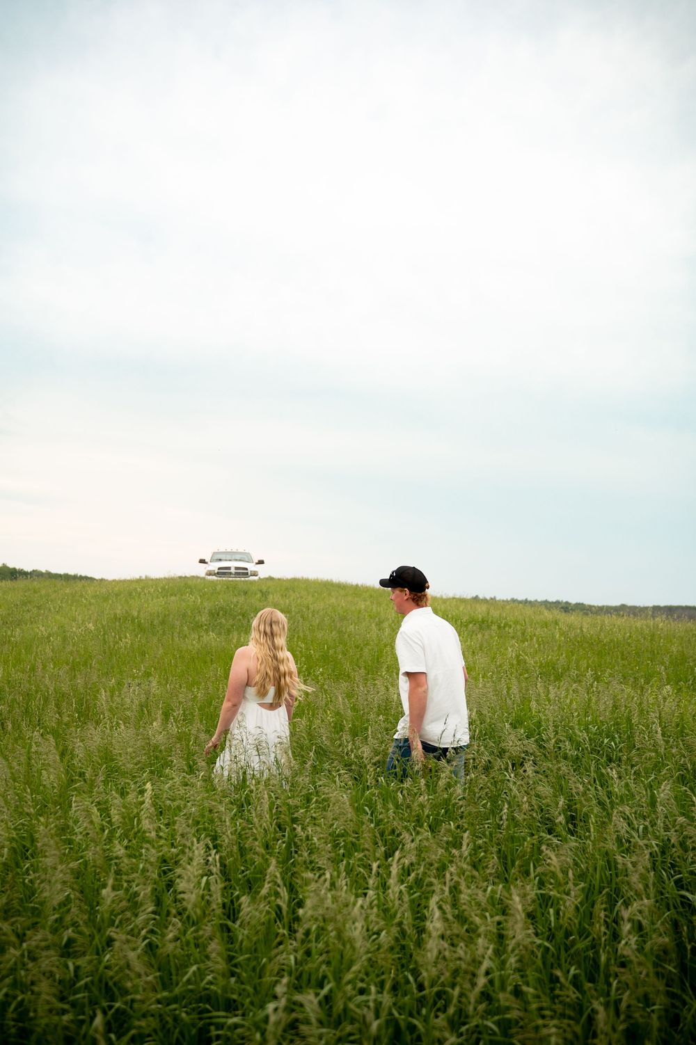 Couple walking through tall green grass field toward a white pickup truck on the horizon.