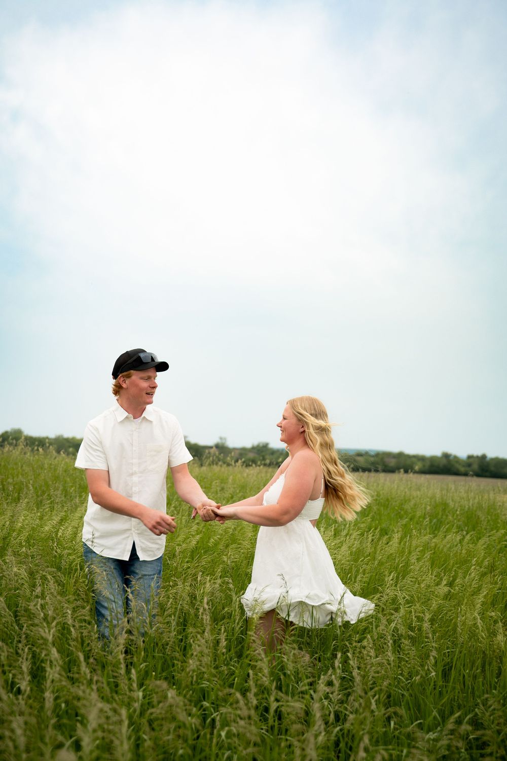 Couple holding hands and dancing together in a tall green grass field, woman in white sundress, man in jeans and cap.