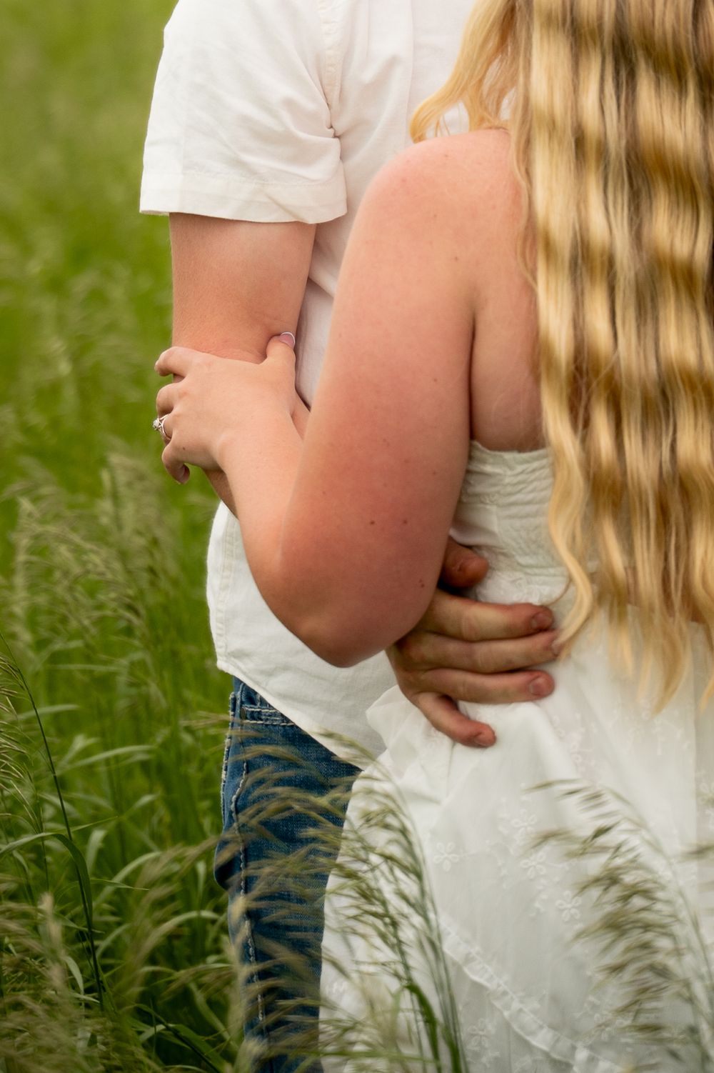 Couple embracing in a green field, woman in white dress with blonde hair, man in white shirt and jeans.