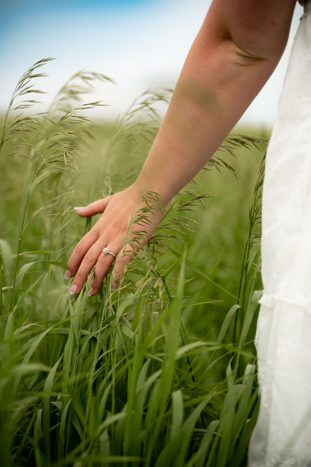 A bride in white dress gently runs her hand through tall green grass, wearing a silver ring, with blue sky background.