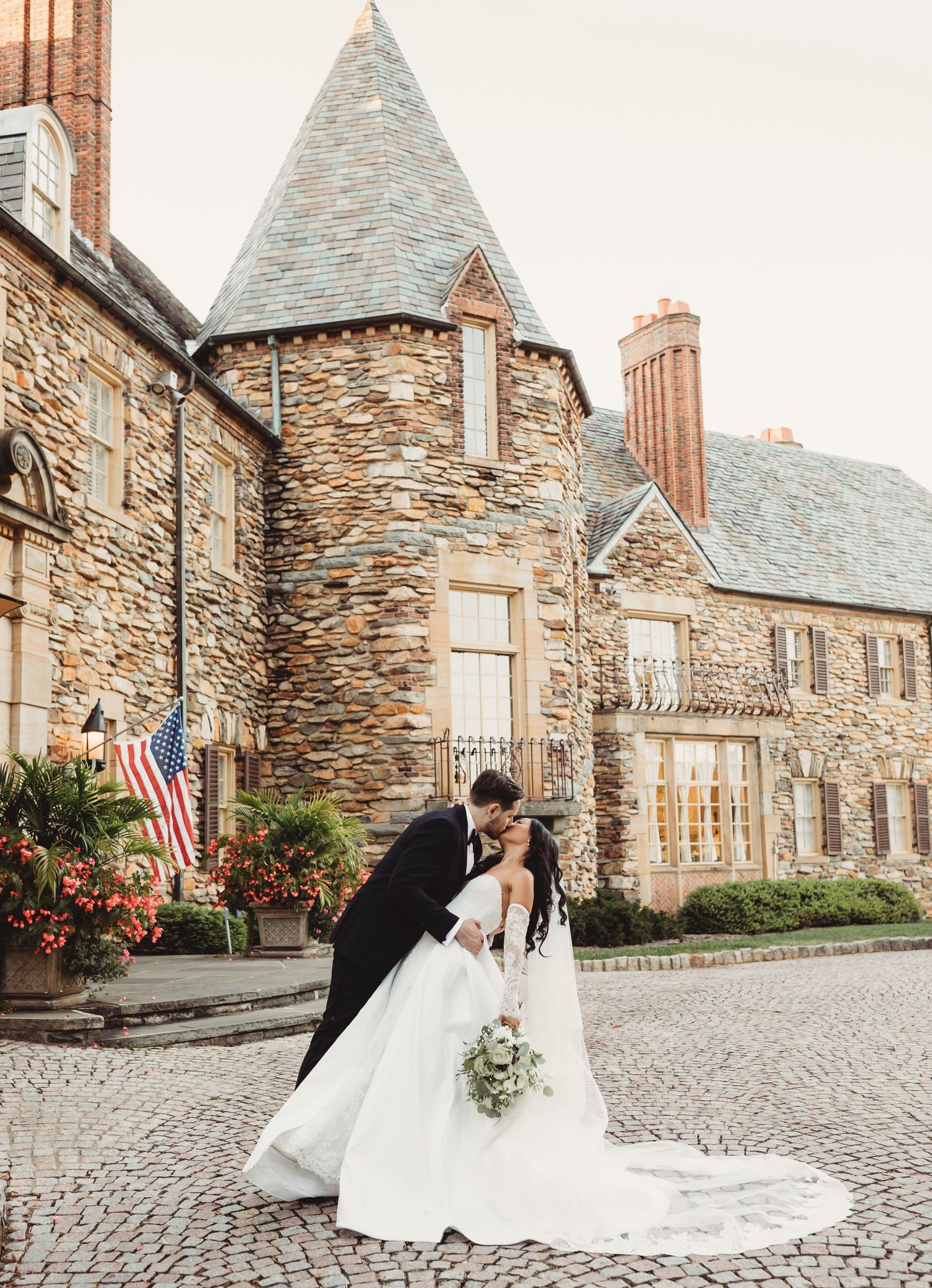 Bride and groom share a romantic dip kiss on cobblestone path before a grand stone castle wedding venue.