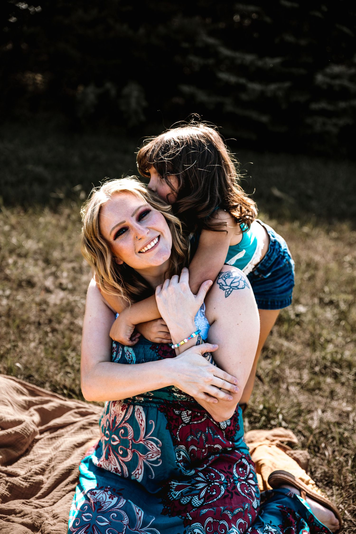 A smiling blonde woman in a floral dress being hugged from behind by a child outdoors on a blanket.
