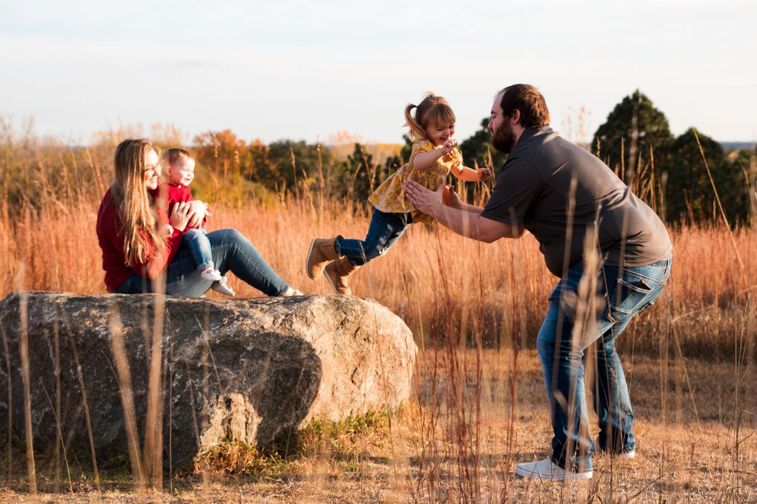 Family enjoying outdoor time together, with parents and two young children playing on a large rock in a field.