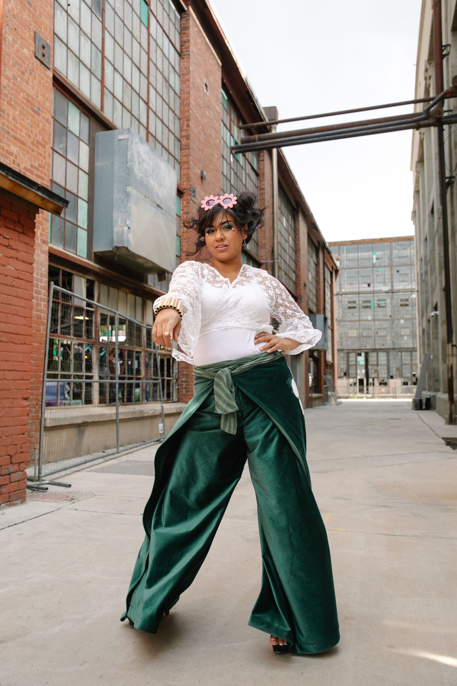 Woman in white lace top and wide-leg green velvet pants poses outdoors near brick buildings.