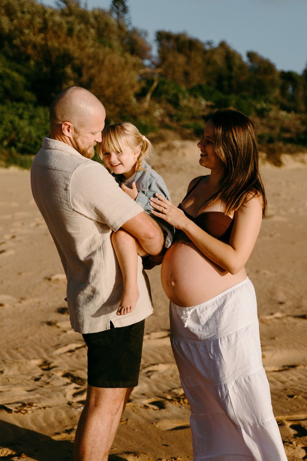 Pregnant woman in white skirt and man holding toddler share tender family moment on sandy beach at golden hour.