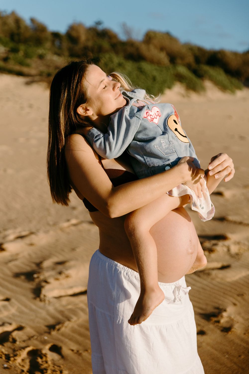 Pregnant woman in white skirt holding baby on sandy beach, sharing a joyful moment in warm sunlight.