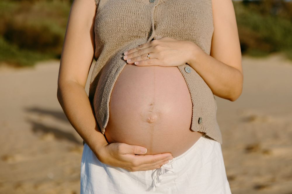 Pregnant woman in beige top and white skirt cradling her bare baby bump outdoors in natural light.
