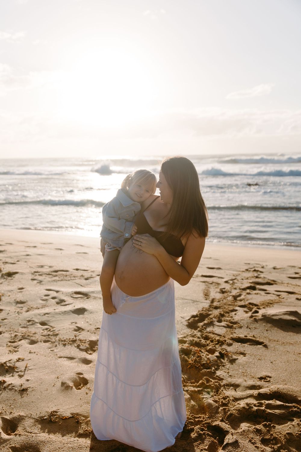 Pregnant woman in white skirt holding toddler on sunny beach with ocean waves.