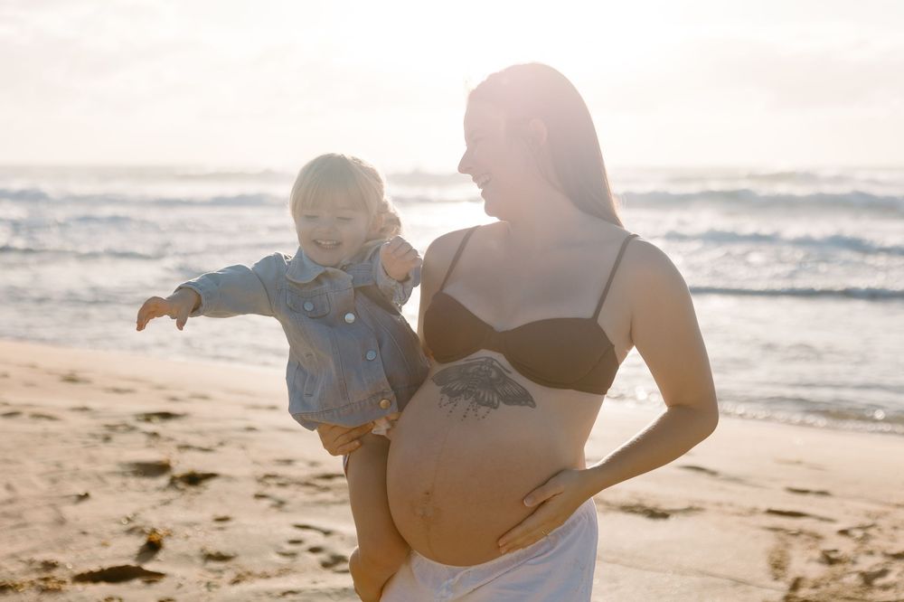 Pregnant woman in black bikini top holds toddler on sunny beach, waves in background.