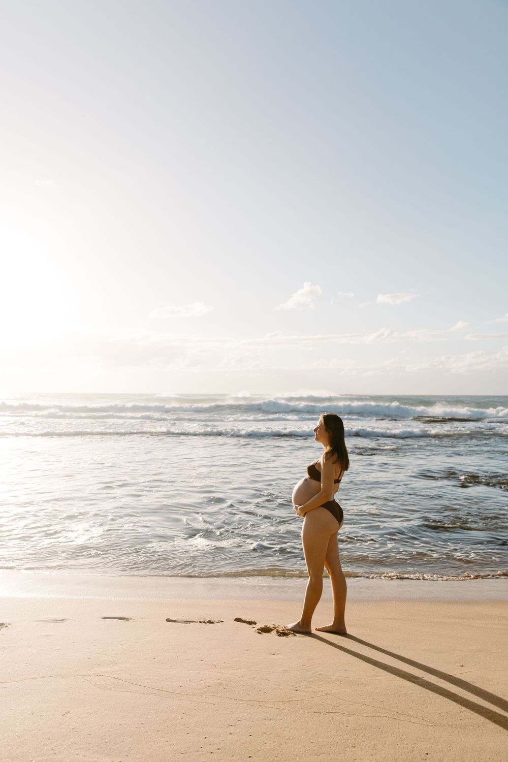 Pregnant woman in bikini cradling belly on sandy beach at golden hour.