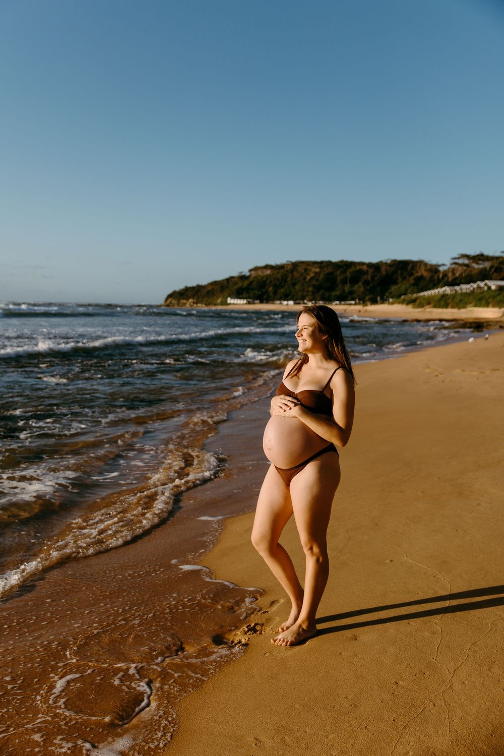 Pregnant woman in brown bikini standing on sandy beach shore, smiling and cradling her baby bump on a sunny day.
