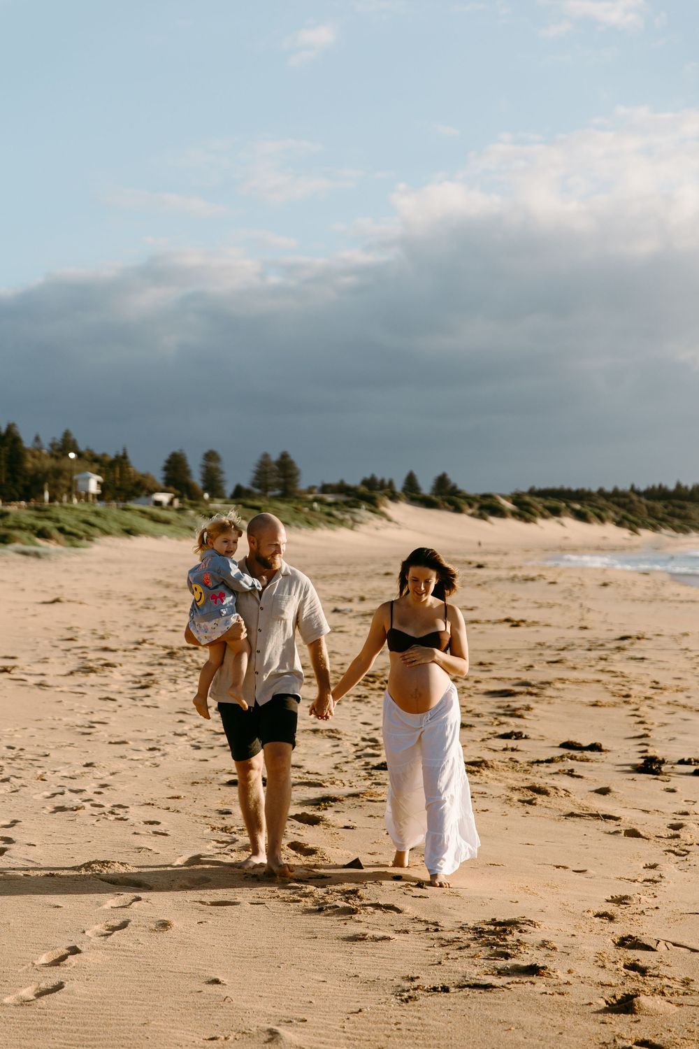 Pregnant woman in bikini top and white skirt walks on beach with partner holding toddler on sunny day.