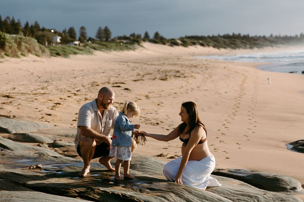 Family of three enjoying a beach day on rocky shores, with a toddler held between parents against a scenic sandy coastline.
