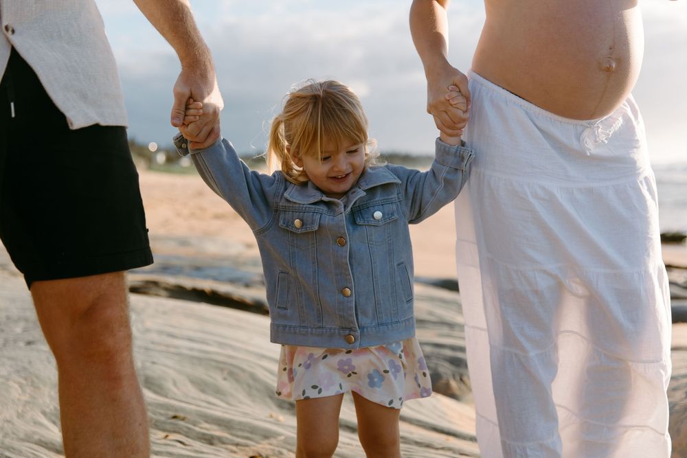 Toddler girl in denim jacket holds parents hands on sandy beach, smiling joyfully during golden hour family photo.