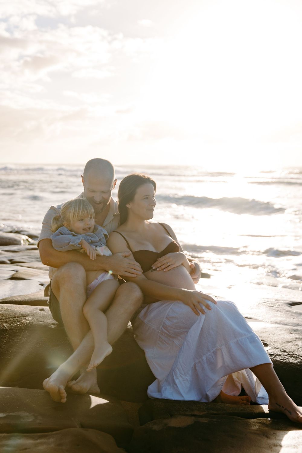 Pregnant woman and man sit on beach rocks at sunset, cradling their toddler in a tender family moment.