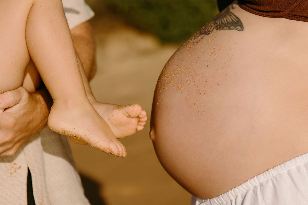 Pregnant woman with butterfly tattoo stands beside a child's bare feet held by another person outdoors.