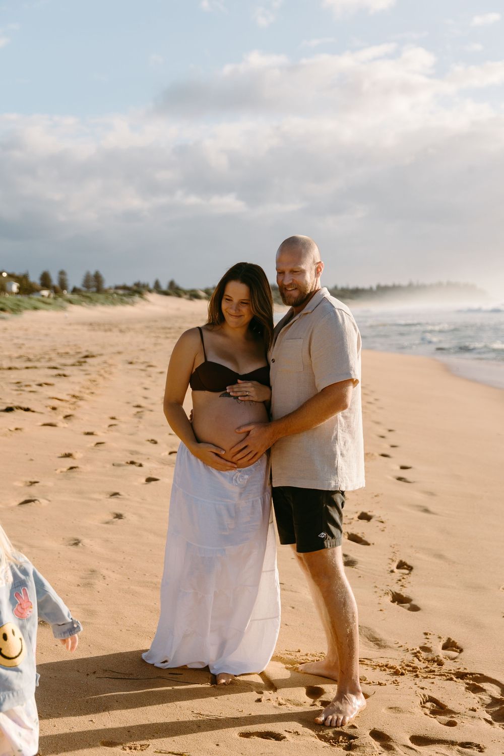 Pregnant couple embracing on sandy beach at golden hour, woman in black bikini top and white skirt, man in grey shirt.