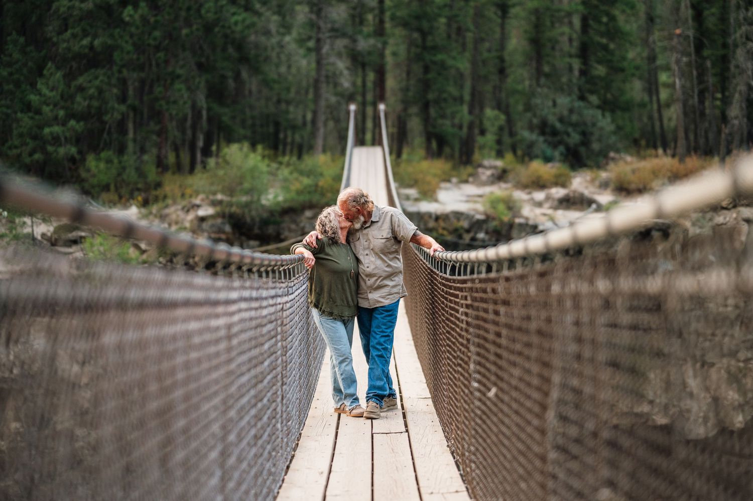Two people walk arm-in-arm across a wooden suspension bridge surrounded by lush forest trees.