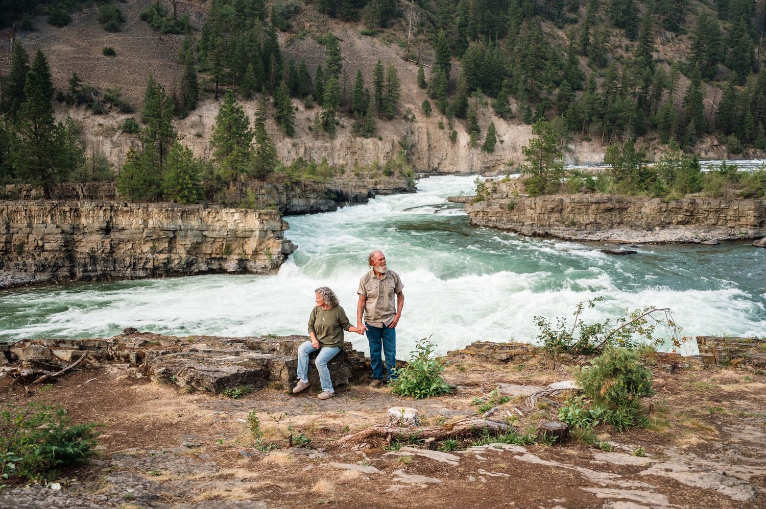 Two people standing on rocky cliffs overlooking a rushing river surrounded by forested mountains.