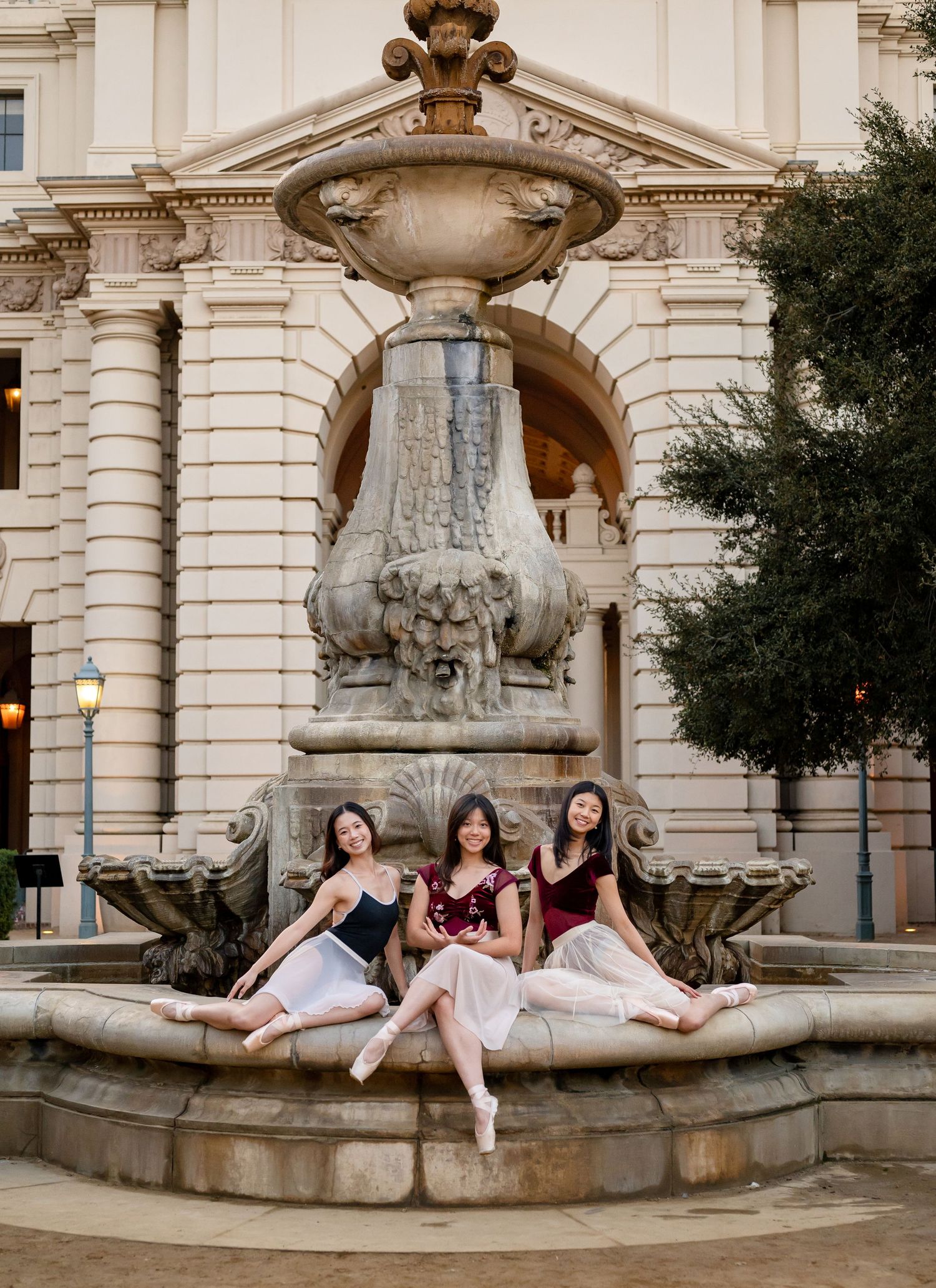 Three ballet dancers pose gracefully on an ornate stone fountain in front of a classical building.