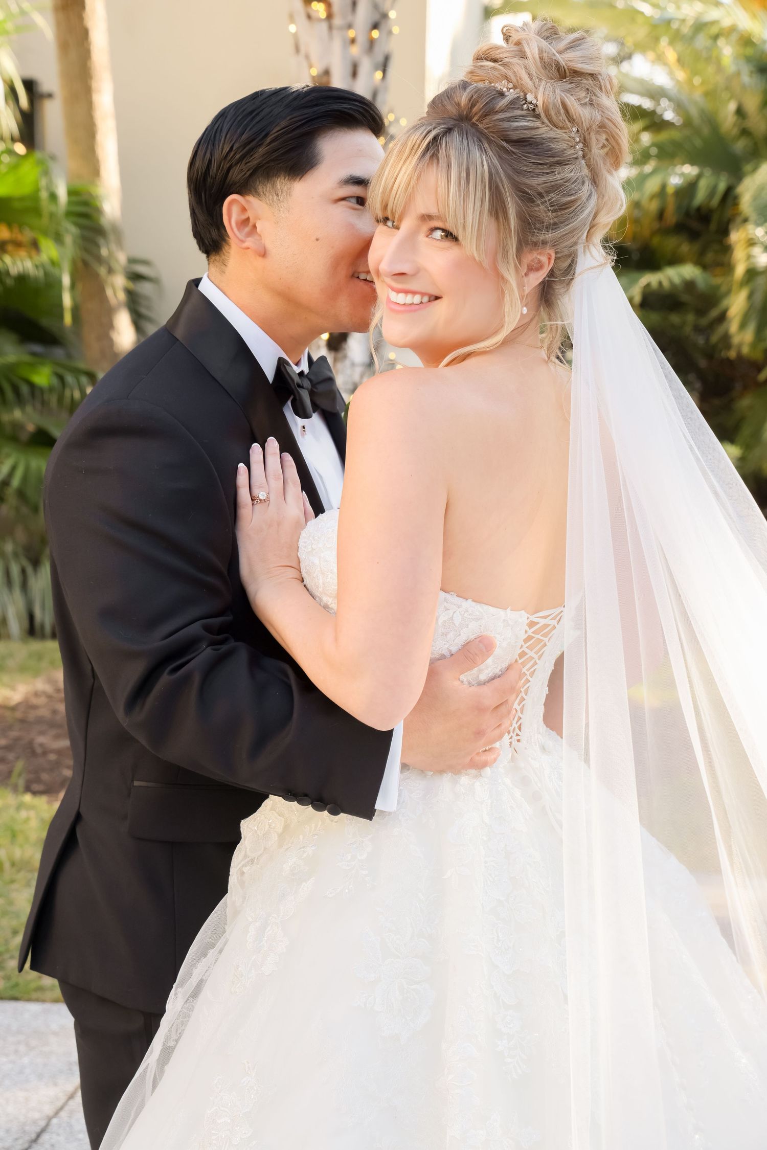 Groom in black tuxedo kissing bride in white strapless gown and veil outdoors at wedding ceremony.