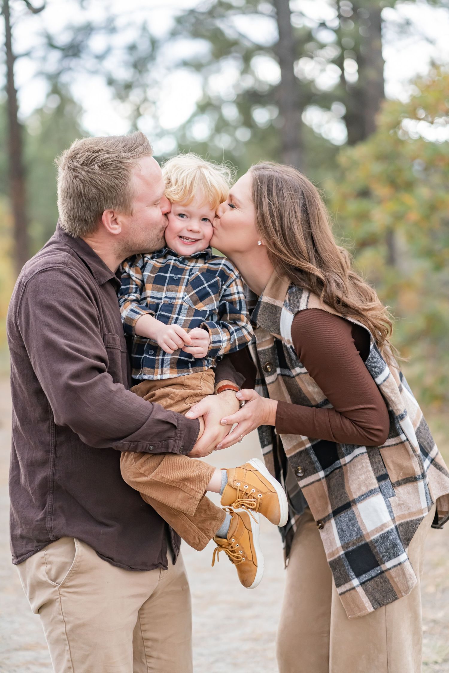 Parents kissing their smiling toddler boy on the cheeks outdoors in an autumn forest setting.