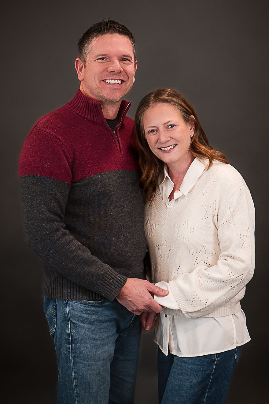 Smiling couple posing together in sweaters against a dark studio background, man in burgundy and gray, woman in white.