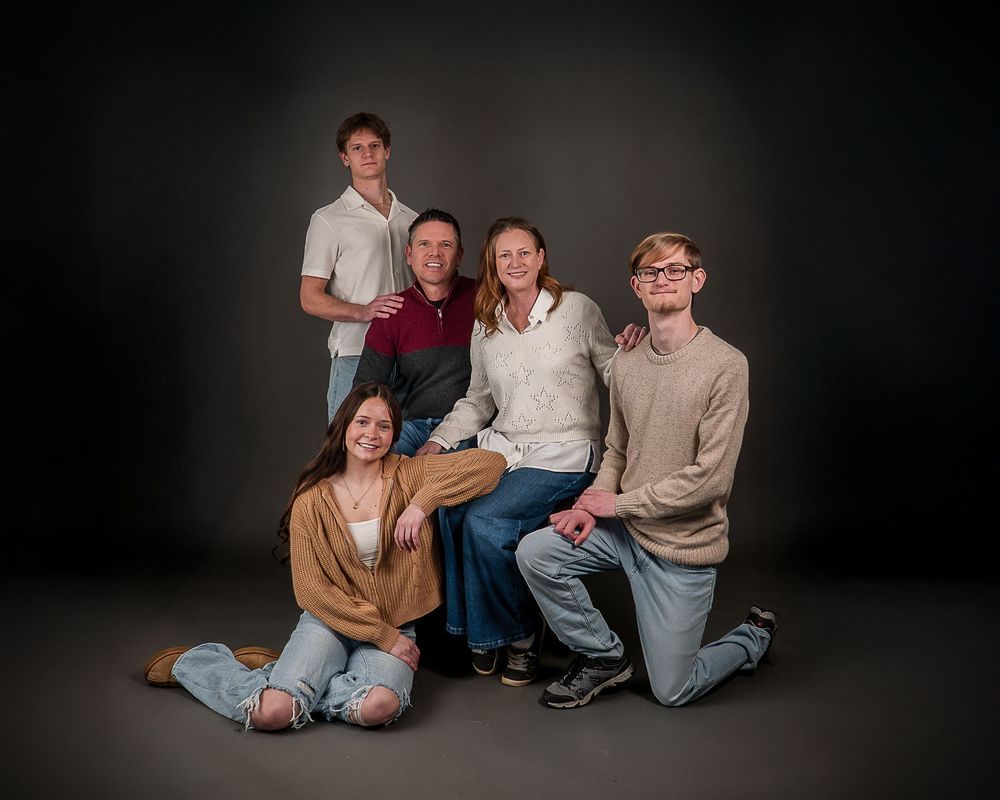 Family of five posing together in a studio portrait, with dark background, two adults seated and three teens around them.