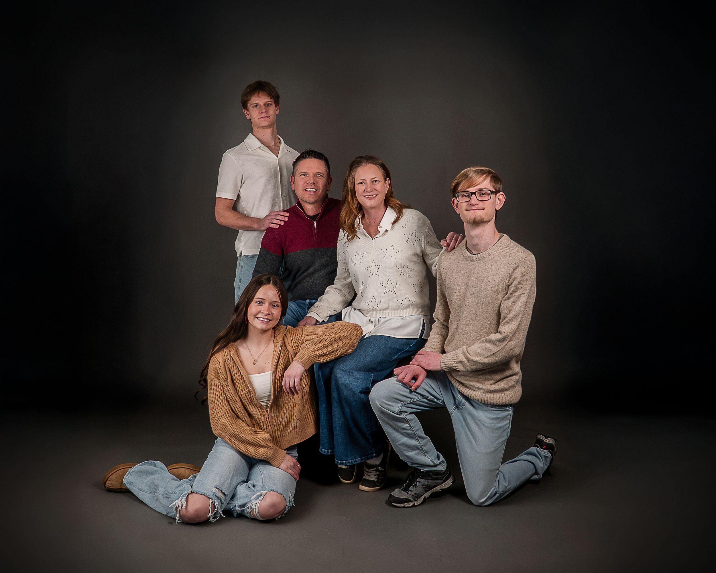 Family of five posing together in a studio portrait, with dark background, two adults seated and three teens around them.