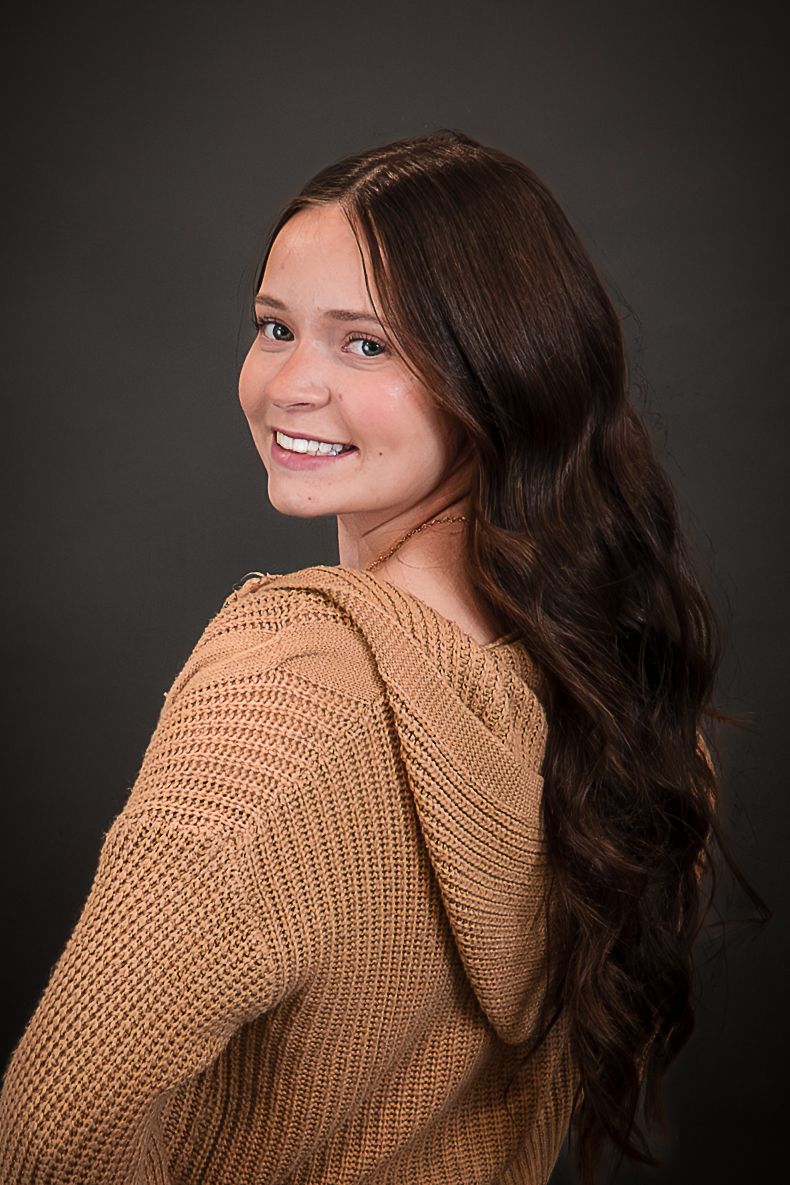 Young woman with long wavy brown hair smiles over shoulder wearing a cozy tan knit sweater against dark background.