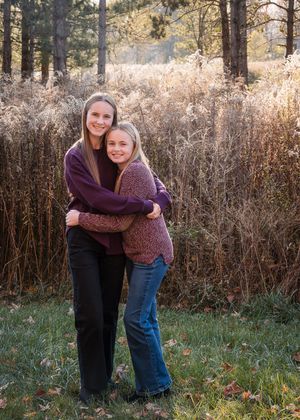 Two girls hugging and smiling outdoors in autumn, wearing purple tops and jeans near tall golden grasses and trees.